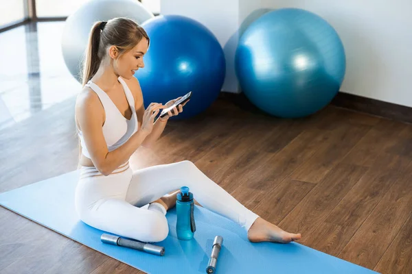 身穿白色运动服的快乐女孩坐在瑜伽垫上 手里拿着智能手机 — 图库照片
