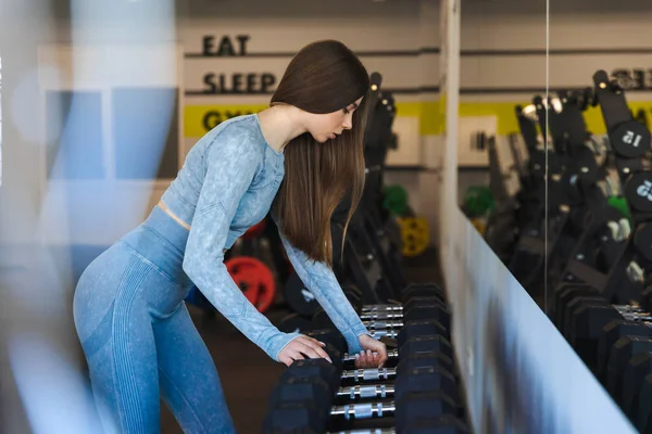 Mavi eşofmanlı kız spor salonundaki raftan bir dambıl alıyor.
