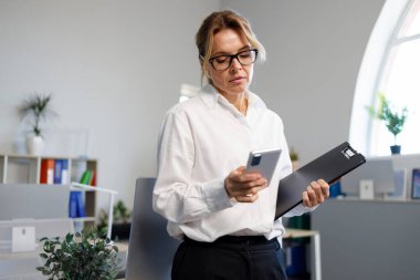 Ofiste beyaz gömlekli ve gözlüklü çekici bir iş kadını akıllı telefon kullanıyor.