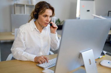 Kulaklıklı kadın çağrı merkezi çalışanı işyerinde oturuyor ve bilgisayar başında çalışıyor.