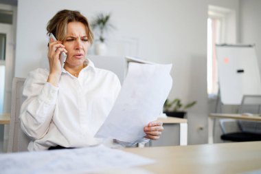 Belgelere bakarken akıllı telefondan konuşan ciddi yüzlü orta yaşlı bir iş kadını.