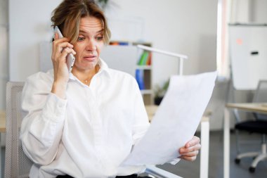 Öfkeli kadın ofis çalışanı belgelere bakıyor ve akıllı telefondan konuşuyor.