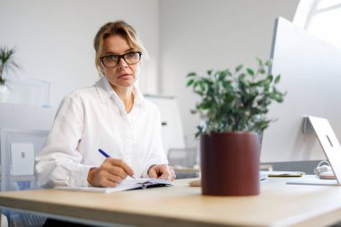 Olgun kadın ofis çalışanı, iş yerinde otururken bir deftere yazar.