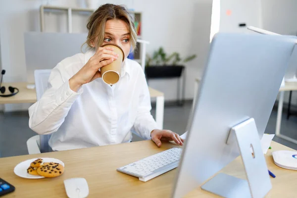 Concentrated at work business woman office worker in a hurry drinking coffee and eating cookie while sitting at the computer