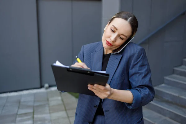 美しいビジネス女性はクリップボードに何かを書き 路上で電話で話します — ストック写真