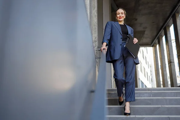 Pretty business woman with clipboard folder walks down the stairs near the office building and looks at the camera