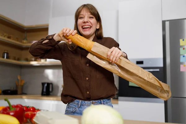 Cheerful girl takes out a french baguette from paper package while cooking in the kitchen