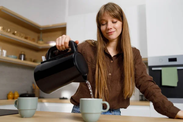 Young woman pouring hot water from electric kettle into mug in kitchen. Girl making tea