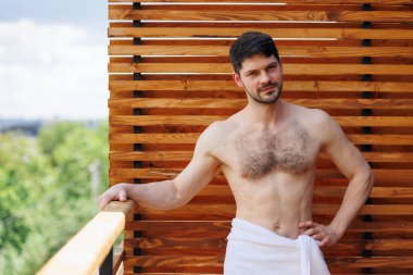 Bir kaplıca otelinin terasında dururken havluyla poz veren çıplak çekici bir adam.