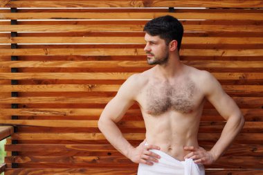 Havlu içinde çekici bir adam, kaplıcadaki ahşap bir duvarın arkasında poz veriyor.