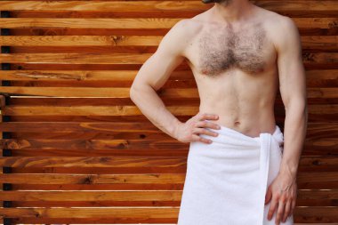 Havlu içinde kaslı vücudu olan bir adam ahşap bir duvarın arka planında poz verir.