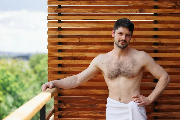 Muscular man in towel posing on terrace of spa resort holding hand on railing
