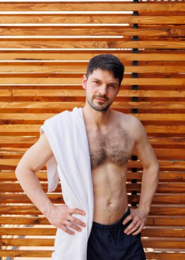 Çekici bir adam, çıplak bir gövdesi ve tahta duvarın arkasında bir havlusu var.