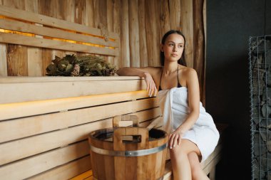 Beyaz havluya sarılı bir kadın saunada bankta oturuyor.