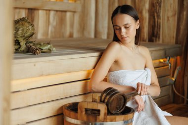 Saunadaki kız daha fazla buhar yapmak için kovadan su topluyor.