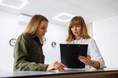 Medical receptionist gives forms to fill to female patient who  standing at reception counter in modern hospital. Woman came for a appointment with a doctor