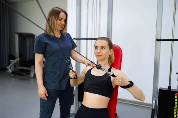 Female rehabilitologist helping woman working out on pull cable machine. Fitness girl is engaged in the gym with a physiotherapist