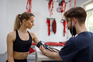 Athletic woman undergoing medical examination in rehabilitation clinic. Doctor measures the arterial pressure of a girl after physical exercise