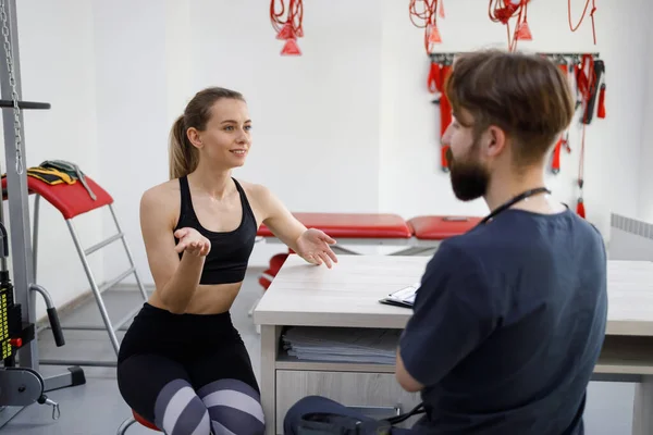 Female patient in a rehabilitation clinic tells a sports doctor about her health problems. Woman consulting with physiotherapist about rehabilitation after injury