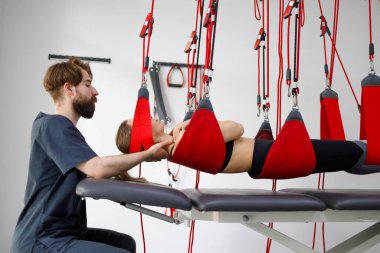 Physiotherapist assists female patient undergoing rehabilitative physiotherapy on a suspension rope system. Modern clinic using advanced rehabilitating therapy