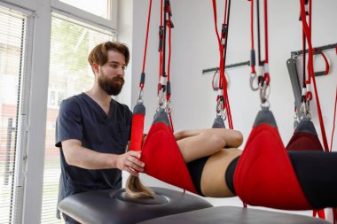 Physiotherapist assists female patient undergoing rehabilitative physiotherapy on a suspension rope system. Modern clinic using advanced rehabilitating therapy