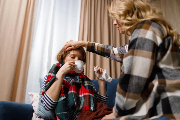 Mature mother takes care of her daughter who is sick with the flu, gives her tea and checks the temperature by placing her palm on her forehead