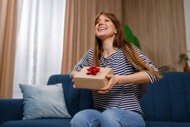 Oturma odasındaki kanepede otururken hediye kutusunu tutan keyifli genç kadın.