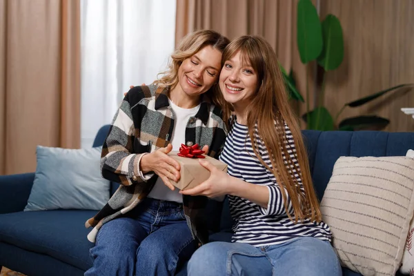 Adult woman makes an unexpected surprise to her middle aged mother, gives her a gift box while sitting on the sofa in apartment