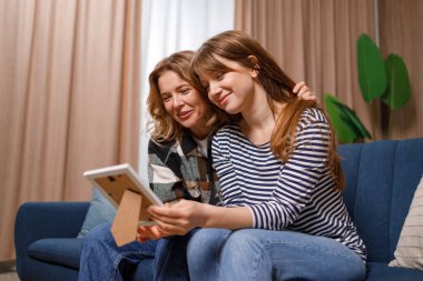 Olgun bir beyaz kadın, kızıyla birlikte kanepede otururken çerçevedeki nostaljik bir fotoğrafa bakar.