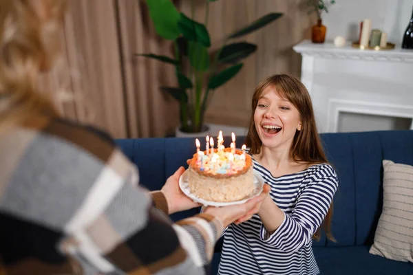 Surprised young woman takes a birthday cake with burning candles from her mother. Make a wish and blow out the candles