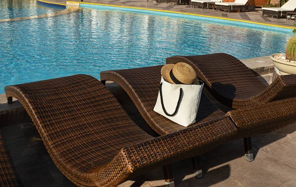 Siyah saplı ve hasır şapkalı beyaz çanta kahverengi güneşli yataklarda insanlar olmadan oturuyor. Sabah ıssız bir havuz manzarası.