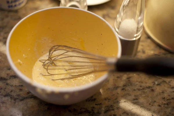 Metal whisk in a mixing bowl with whisked egg on a kitchen table reading for baking