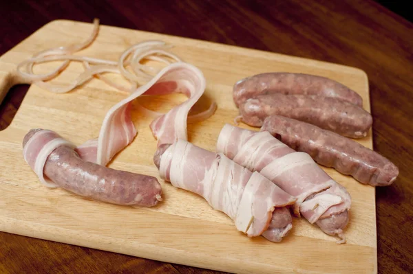 It wouldn\'t be Christmas: Preparing bacon rolls on a wooden board in the kitchen with spicy sausages and rashers of smoked bacon for the wrapper