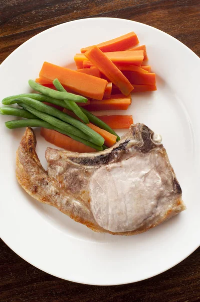 Grilled pork cutlets with crispy fat served with fresh sliced carrots and runner beans on a white plate, overhead view
