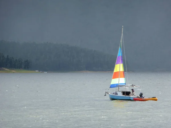 Small sailboat with a single colourful sail towing a dinghy offshore on a misty cold day with the coastline visible in the distance