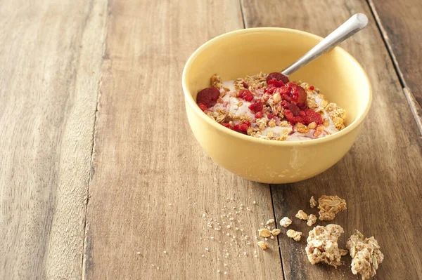 Spilled granola chunks next to partially full cereal bowl that includes raspberry fruit over wooden table with copy space