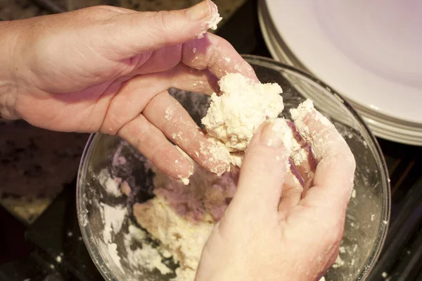 Hands shaping course dough into dumplings over glass bowl beside stack of white plates