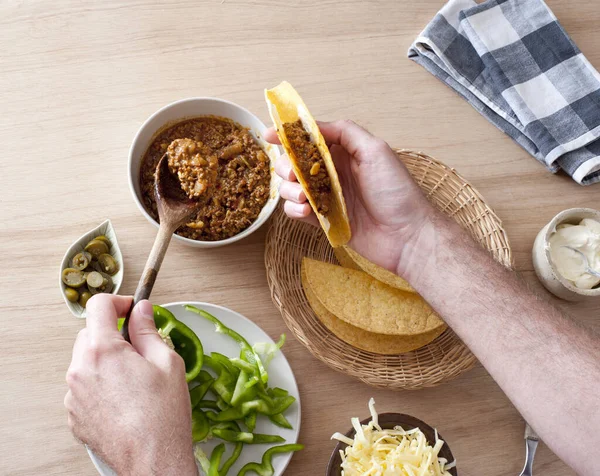 First person perspective view of hands filling a taco shell with ground meat beside green and hot peppers, lettuce and sour cream