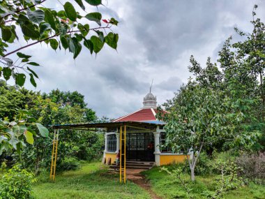 Beyaz ve açık sarı renkli piramit şekilli çatıya boyanmış küçük yuvarlak Hindu tapınağının stok fotoğrafı. Gulbarga 'daki yeşil ağaçlar ve bitkilerle çevrili tapınağın önünde küçük bir barınak.