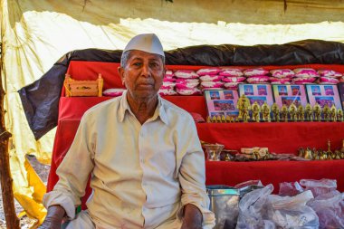 Kolhapur, Hindistan - 31 Ocak 2019; 50-60 yaş arası, geleneksel beyaz gömlek, pantolon ve kep giyen Hintli dedesinin dükkanında ibadet eşyaları satarken çekilmiş resmi. Arkaplanda küçük bir dükkan