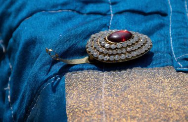 Broşun ortasında küçük beyaz elmas, koyu kırmızı renkli taşla süslenmiş güzel yuvarlak broş resmi. Arkaplanda parlak güneş ışığı altında çekilmiş mavi renk kumaş.