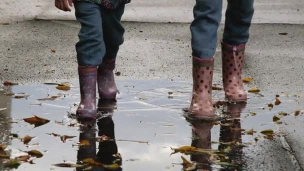 Dos niñas con botas de goma caminan sobre asfalto húmedo cubierto de charcos y hojas caídas. Niños y otoño. Vídeo en cámara lenta Full HD — Vídeo de stock