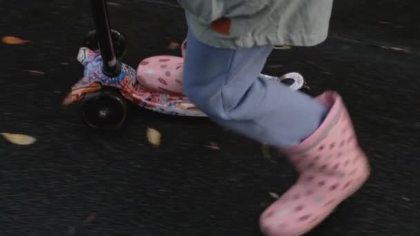 Kind meisje in rubberen laarzen rijdt op een nat asfalt bedekt met plassen en gevallen bladeren op een scooter. Full HD slow motion close-up video. — Stockvideo