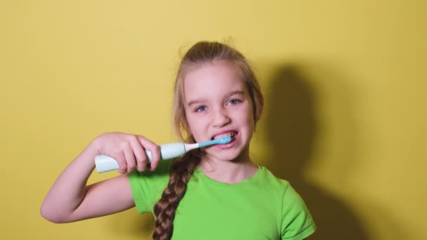 Tonårstjej borstar tänderna isolerade på ljusgul bakgrund. Barnflicka i grön t-shirt använder en elektrisk tandborste för att borsta tänderna — Stockvideo