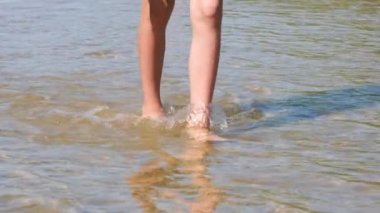 Sahildeki sığ suda ıslak kumda yürüyen neşeli çocuk bacaklarına yakından bak. Olumlu duygular hisset. Tam HD yavaş çekim videosu. Yaz tatili kavramı