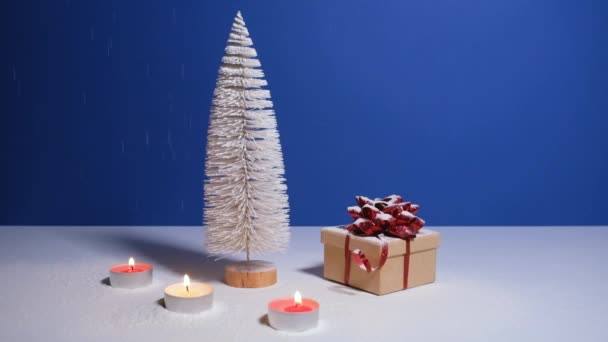 Belle bannière vidéo de Noël ou Nouvel An avec espace de copie. Jouet sapin de Noël, boîte cadeau avec arc rouge et bougies allumées sur fond bleu avec neige tombant sur le fond. — Video