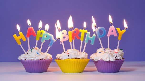 Feliz cumpleaños Inscripción hecha de velas de colores ardientes en sabrosos cupcakes aislados sobre fondo púrpura. Feliz cumpleaños banner de vídeo, cámara lenta — Vídeo de stock
