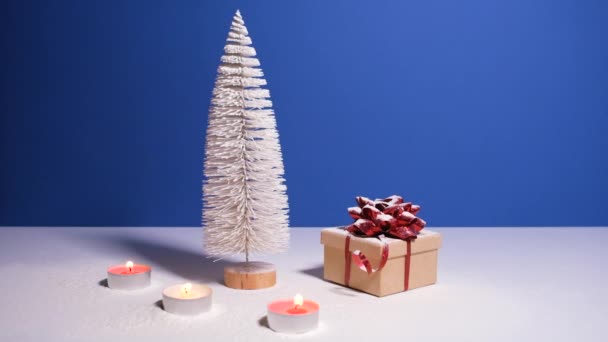 Hermoso banner de Navidad o Año Nuevo con espacio para copiar. Árbol de Navidad de juguete, caja de regalo con lazo rojo y velas ardientes sobre fondo azul con nieve cayendo en el fondo. — Vídeos de Stock