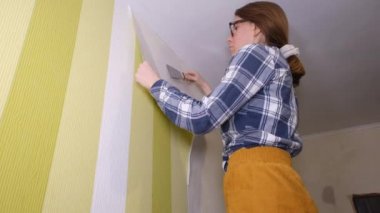 Elinde spatulası olan genç bir kadın tasarımcı merdiven üzerinde duruyor ve duvardaki eski duvar kağıdını yırtıyor. Daire yenileme konsepti
