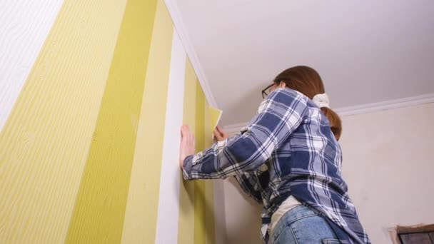 Una joven diseñadora con una espátula en la mano se para en una escalera y arranca el viejo papel pintado de las paredes. Concepto de renovación de apartamentos — Vídeo de stock